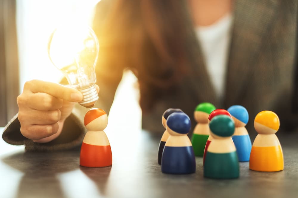 Close up of a person holding a lit lightbulb near colorful wooden figures on a table symbolizing lead generation services.